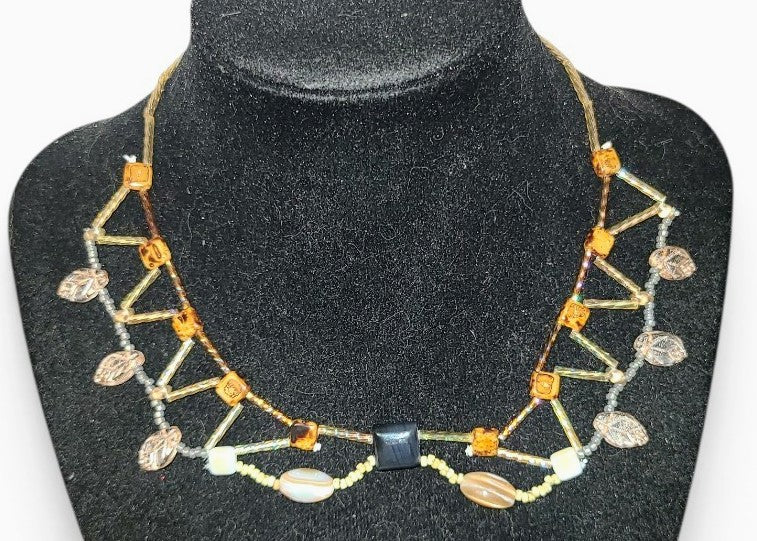 Architectural beaded necklace with square black onyx center stone, shell ovals, amber glass cube beads, and champagne leaf drops on a dark background.