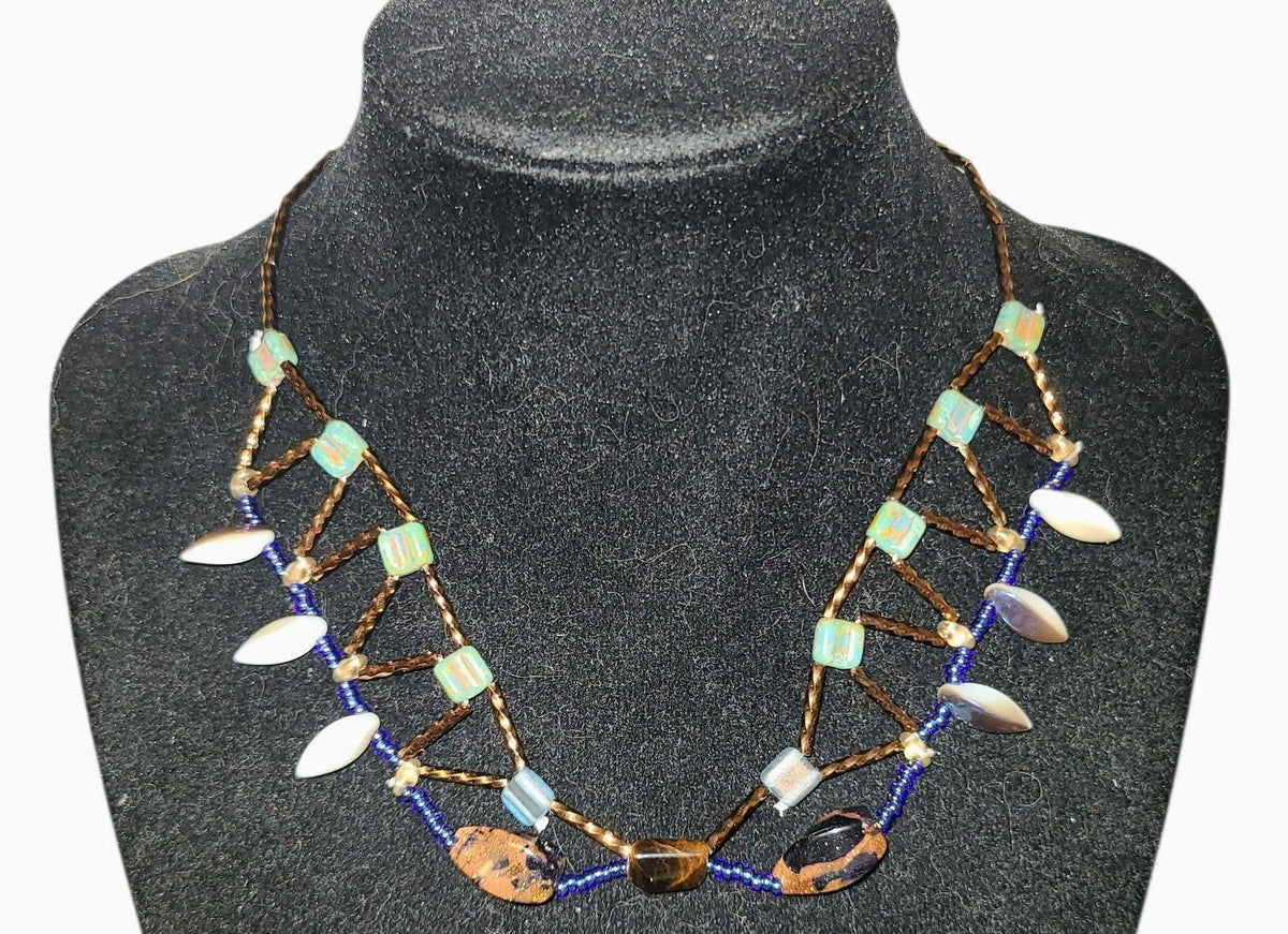 Architectural beaded necklace with South African tiger eye center stone, goldstone oval, cobalt blue seed beads, and iridescent glass drops on a dark background.