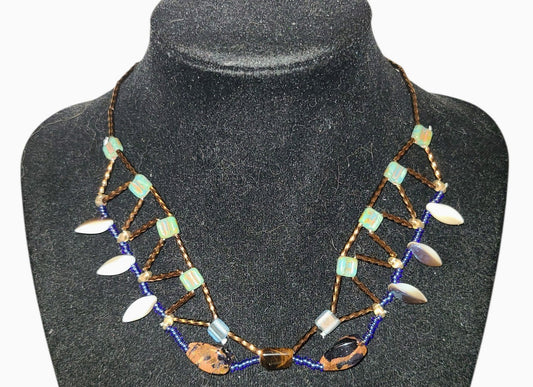 Architectural beaded necklace with South African tiger eye center stone, goldstone oval, cobalt blue seed beads, and iridescent glass drops on a dark background.